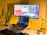 A cozy home office setup with a laptop on a stand displaying code on the screen, accompanied by an external monitor showing colorful graphics. The background is a warm yellow wall, and the desk features a keyboard, a black mouse pad, and two potted plants. A small plush toy is also present beside the plants, contributing to a relaxed atmosphere.