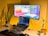 A cozy home office setup with a laptop on a stand displaying code on the screen, accompanied by an external monitor showing colorful graphics. The background is a warm yellow wall, and the desk features a keyboard, a black mouse pad, and two potted plants. A small plush toy is also present beside the plants, contributing to a relaxed atmosphere.