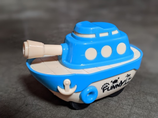 A small plastic toy that resembles a cartoonish battleship or boat. The toy is primarily blue with cream-colored accents, including a cannon on the front and several circular windows. It has the word 'FUNNY' printed on the side, accompanied by the image of a crab and palm trees.