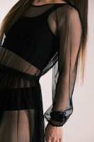 A person wearing a sheer black dress with long sleeves. The fabric appears translucent, revealing another layer underneath. The style is elegant and modern.