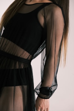 A person wearing a sheer black dress with long sleeves. The fabric appears translucent, revealing another layer underneath. The style is elegant and modern.