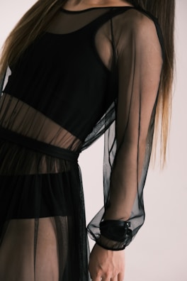 A person wearing a sheer black dress with long sleeves. The fabric appears translucent, revealing another layer underneath. The style is elegant and modern.