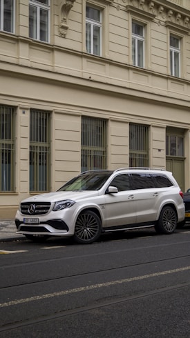 A white Mercedes SUV is parked on a city street beside a beige, ornate building with several tall windows that have metal bars.