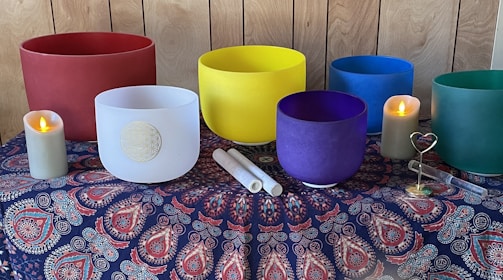 A collection of colorful singing bowls arranged on a patterned fabric tablecloth, with two lit candles on either side. The singing bowls are in hues of red, white, yellow, blue, purple, and green. Two mallets are placed in front of the white bowl. The tablecloth features intricate patterns in red, blue, and white on a dark background. The setting appears to be calm and harmonious.
