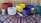 A collection of colorful singing bowls arranged on a patterned fabric tablecloth, with two lit candles on either side. The singing bowls are in hues of red, white, yellow, blue, purple, and green. Two mallets are placed in front of the white bowl. The tablecloth features intricate patterns in red, blue, and white on a dark background. The setting appears to be calm and harmonious.