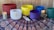 A collection of colorful singing bowls arranged on a patterned fabric tablecloth, with two lit candles on either side. The singing bowls are in hues of red, white, yellow, blue, purple, and green. Two mallets are placed in front of the white bowl. The tablecloth features intricate patterns in red, blue, and white on a dark background. The setting appears to be calm and harmonious.
