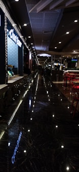 A dimly lit corridor in a modern establishment with reflective marble flooring, overhead lights, tables with red chairs on one side, and a sign reading 'Chaat Bazaar' on the other. The ceiling features intricate design with recessed lighting. The atmosphere appears elegant and contemporary.