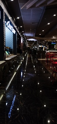 A dimly lit corridor in a modern establishment with reflective marble flooring, overhead lights, tables with red chairs on one side, and a sign reading 