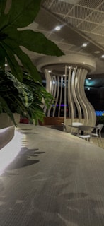 A modern indoor space featuring a curved, wooden table surface with reflections. In the background, there is a unique structural element resembling a large vase or column with wavy vertical slats. The ceiling has a grid pattern with recessed lighting. Green foliage adds a touch of nature to the scene.