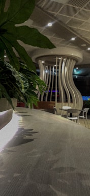 A modern indoor space featuring a curved, wooden table surface with reflections. In the background, there is a unique structural element resembling a large vase or column with wavy vertical slats. The ceiling has a grid pattern with recessed lighting. Green foliage adds a touch of nature to the scene.