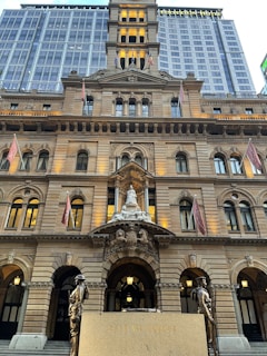 A grand, multi-story historic building with intricate architectural details, including arched windows and ornate sculptures. Two flags are displayed, and there is a bronze statue in front of a memorial with the words 'Lest We Forget' inscribed.