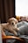 A golden retriever puppy is lying comfortably on a gray, patterned blanket draped over a chair. In the background, a window sill holds a yellow house-shaped decoration and two mugs.