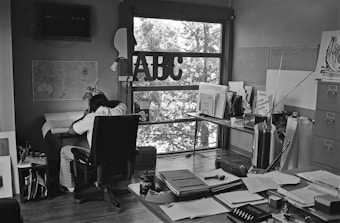 A workspace with a person sitting at a small drafting table, surrounded by various papers and office supplies. A window with large lettering allows light to pour in, casting shadows of trees outside into the room. The space includes a map on the wall, filing cabinets, and a cluttered desk with documents and a can of Folger's coffee.