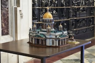 A detailed architectural model of a historic building with a prominent golden dome and intricate design features, displayed on a wooden table. The surrounding area includes marble and ornate carved elements, adding to the sophisticated atmosphere.