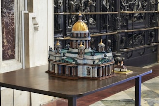 A detailed architectural model of a historic building with a prominent golden dome and intricate design features, displayed on a wooden table. The surrounding area includes marble and ornate carved elements, adding to the sophisticated atmosphere.