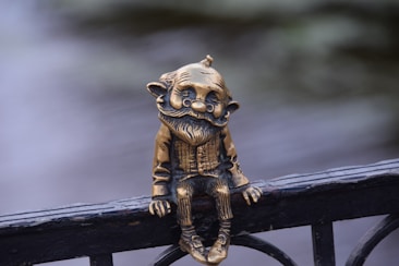 A bronze figurine of a gnome is perched on a railing. The gnome has an exaggerated facial expression with a large mustache and ears. The background is blurred, emphasizing the figurine.