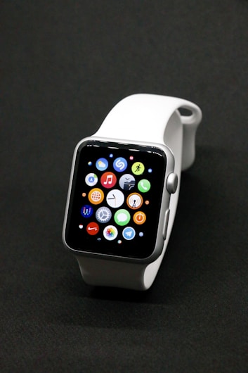 A sleek smartwatch with a silver case and a white band, featuring a vibrant display showing various app icons against a dark background.