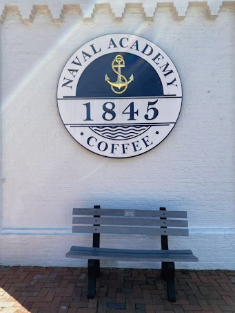 A circular sign mounted on a white brick wall with the words 'Naval Academy Coffee 1845' and an anchor symbol in the center. Below the sign, a gray wooden bench is placed on a red brick pavement.