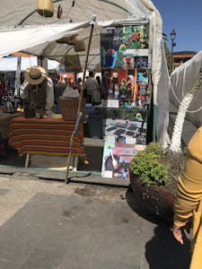 A market stall features a colorful tablecloth and various items for sale, including baskets and a kettle. A person wearing a hat is attending to the stall. There are informational posters with images of coffee beans and processing displayed. The area is covered by a white tent, providing shade.