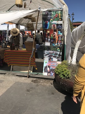 A market stall features a colorful tablecloth and various items for sale, including baskets and a kettle. A person wearing a hat is attending to the stall. There are informational posters with images of coffee beans and processing displayed. The area is covered by a white tent, providing shade.
