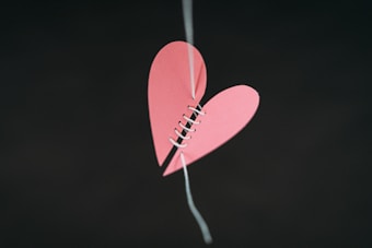 A pink paper heart split in half is sewn back together with white string, hanging against a dark background.