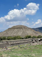 A large ancient pyramid structure surrounded by greenery under a bright blue sky with fluffy clouds. A group of people is gathered near the base of the pyramid, possibly tourists or visitors, exploring the site. The landscape features a mix of natural vegetation and stone architecture.