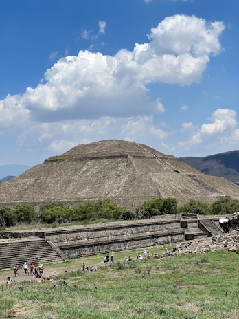 A large ancient pyramid structure surrounded by greenery under a bright blue sky with fluffy clouds. A group of people is gathered near the base of the pyramid, possibly tourists or visitors, exploring the site. The landscape features a mix of natural vegetation and stone architecture.