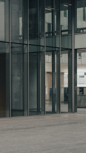 Tall glass windows reflecting an urban exterior, with clean architectural lines and a minimalistic design. The ground is a smooth concrete surface, contributing to a modern and sleek environment.