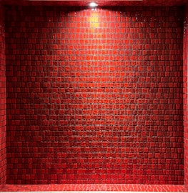 A wall composed of red, square tiles under a bright light, casting subtle shadows. The tiles have a glossy finish and are arranged in a grid-like pattern, creating a textured and uniform surface.
