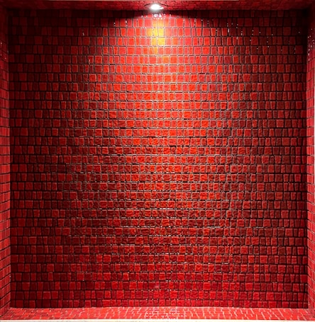A wall composed of red, square tiles under a bright light, casting subtle shadows. The tiles have a glossy finish and are arranged in a grid-like pattern, creating a textured and uniform surface.