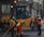 Several construction workers wearing high-visibility safety vests and helmets are actively engaged in roadwork. A large yellow road roller is parked nearby as the workers use shovels and other tools to smooth and prepare the pavement.