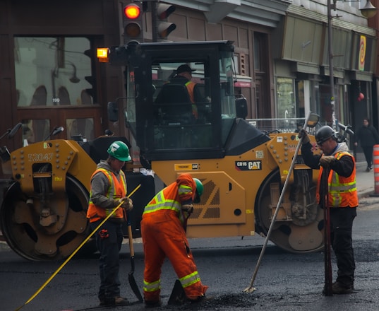 Several construction workers wearing high-visibility safety vests and helmets are actively engaged in roadwork. A large yellow road roller is parked nearby as the workers use shovels and other tools to smooth and prepare the pavement.