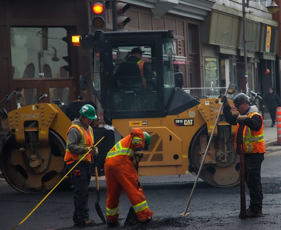 Several construction workers wearing high-visibility safety vests and helmets are actively engaged in roadwork. A large yellow road roller is parked nearby as the workers use shovels and other tools to smooth and prepare the pavement.