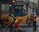 Several construction workers wearing high-visibility safety vests and helmets are actively engaged in roadwork. A large yellow road roller is parked nearby as the workers use shovels and other tools to smooth and prepare the pavement.