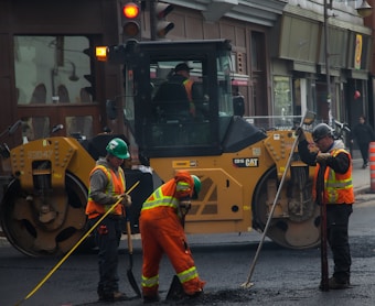 Several construction workers wearing high-visibility safety vests and helmets are actively engaged in roadwork. A large yellow road roller is parked nearby as the workers use shovels and other tools to smooth and prepare the pavement.
