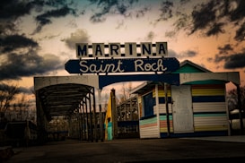 An entrance sign reads 'Marina Saint Roch' with a rustic, colorful booth nearby. The structure has a weathered wooden canopy, leading into a boardwalk or dock area surrounded by leafless trees. The sky is dramatic, with dark clouds contrasted by a warm sunset.