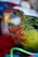A close-up of a colorful parrot with vibrant feathers in shades of red, yellow, and green. The bird has a distinctive curved beak and round, dark eyes. In the background, various colorful objects, including a red thread ball and green string, are slightly blurred.
