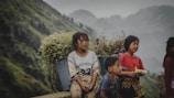 A group of children is seen against a backdrop of lush green hills. One child is sitting, carrying a basket full of wildflowers on their back, while the others stand nearby. Their clothing appears traditional and colorful, reflecting cultural attire.
