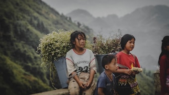 A group of children is seen against a backdrop of lush green hills. One child is sitting, carrying a basket full of wildflowers on their back, while the others stand nearby. Their clothing appears traditional and colorful, reflecting cultural attire.