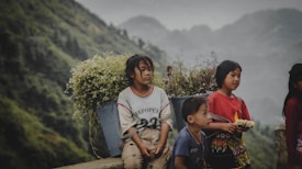 A group of children is seen against a backdrop of lush green hills. One child is sitting, carrying a basket full of wildflowers on their back, while the others stand nearby. Their clothing appears traditional and colorful, reflecting cultural attire.