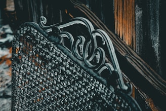 A detailed view of an ornate metal structure featuring intricate scrollwork patterns and a mesh-like texture. The background includes vertical, weathered wooden planks with a warm, contrasting hue.