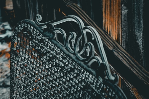 A detailed view of an ornate metal structure featuring intricate scrollwork patterns and a mesh-like texture. The background includes vertical, weathered wooden planks with a warm, contrasting hue.