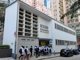 A group of people, mostly wearing school uniforms, are entering a white building labeled 'Bridges Street Market' and 'Hong Kong News-Expo'. The structure is located in an urban area surrounded by tall residential buildings and parked cars. A no U-turn sign is visible in the foreground along with some greenery.