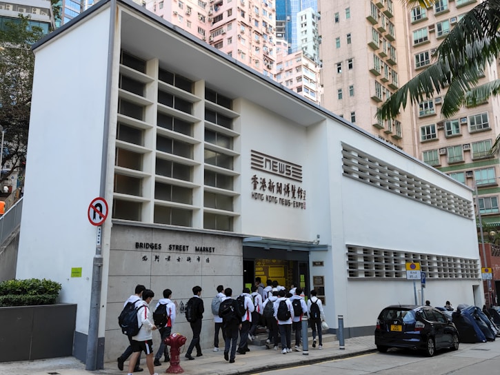 A group of people, mostly wearing school uniforms, are entering a white building labeled 'Bridges Street Market' and 'Hong Kong News-Expo'. The structure is located in an urban area surrounded by tall residential buildings and parked cars. A no U-turn sign is visible in the foreground along with some greenery.