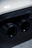Close-up of a car's dual exhaust pipes featuring a glossy carbon fiber finish, with reflections on the surface. The image includes part of the car's rear bumper, which is white, adding a contrast to the dark exhaust.