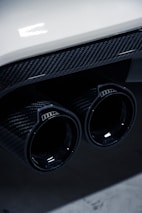 Close-up of a car's dual exhaust pipes featuring a glossy carbon fiber finish, with reflections on the surface. The image includes part of the car's rear bumper, which is white, adding a contrast to the dark exhaust.