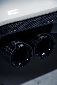 Close-up of a car's dual exhaust pipes featuring a glossy carbon fiber finish, with reflections on the surface. The image includes part of the car's rear bumper, which is white, adding a contrast to the dark exhaust.