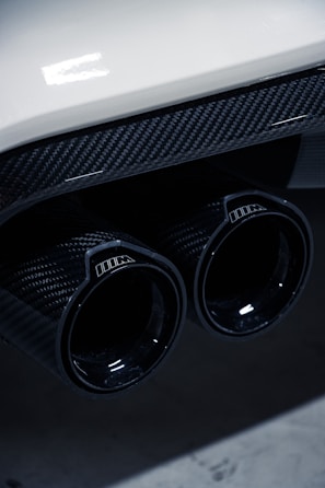 Close-up of a car's dual exhaust pipes featuring a glossy carbon fiber finish, with reflections on the surface. The image includes part of the car's rear bumper, which is white, adding a contrast to the dark exhaust.