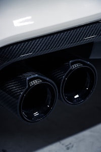 Close-up of a car's dual exhaust pipes featuring a glossy carbon fiber finish, with reflections on the surface. The image includes part of the car's rear bumper, which is white, adding a contrast to the dark exhaust.