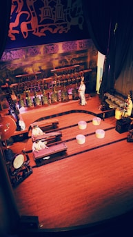 A traditional stage performance with musicians dressed in cultural attire playing various instruments. The setting features ornate decorations and traditional motifs with intricate patterns on the backdrop. Large percussion instruments and string instruments are visible, with performers engaged in a musical presentation.